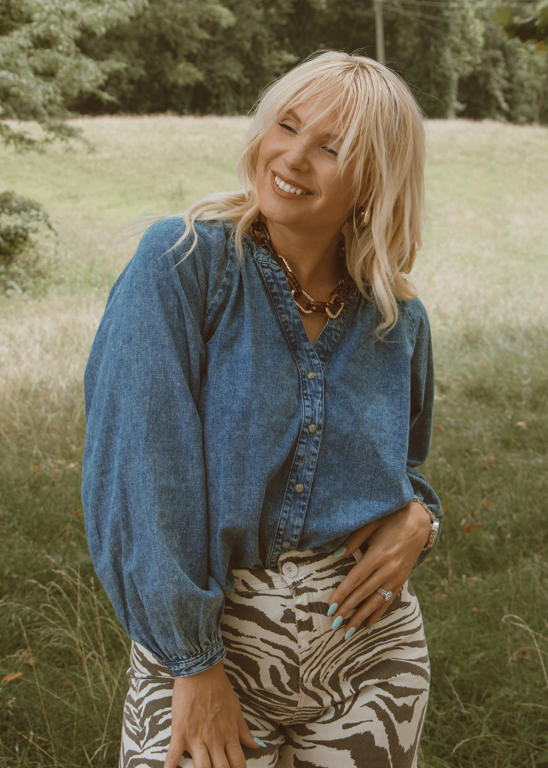 Woman wearing a blue denim shirt and zebra print pants standing in a grassy field.