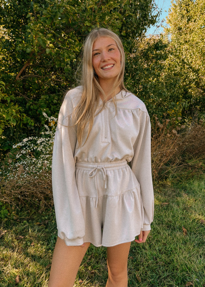 Woman in a beige romper standing outdoors with greenery in the background