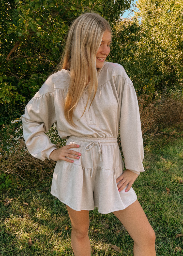 Woman wearing a beige long-sleeve romper standing in a natural setting with greenery.