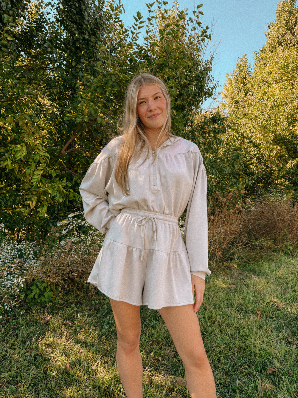 Woman in a light-colored romper standing in a natural setting with trees and grass.