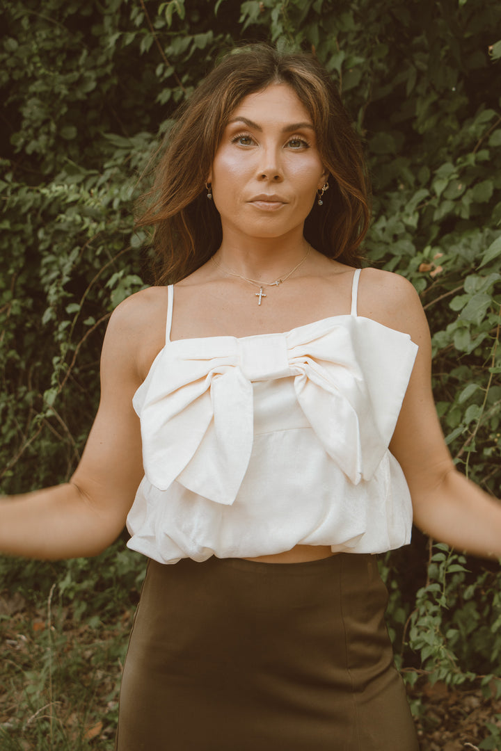 Woman wearing a white top with a bow and brown pants against a green leafy background