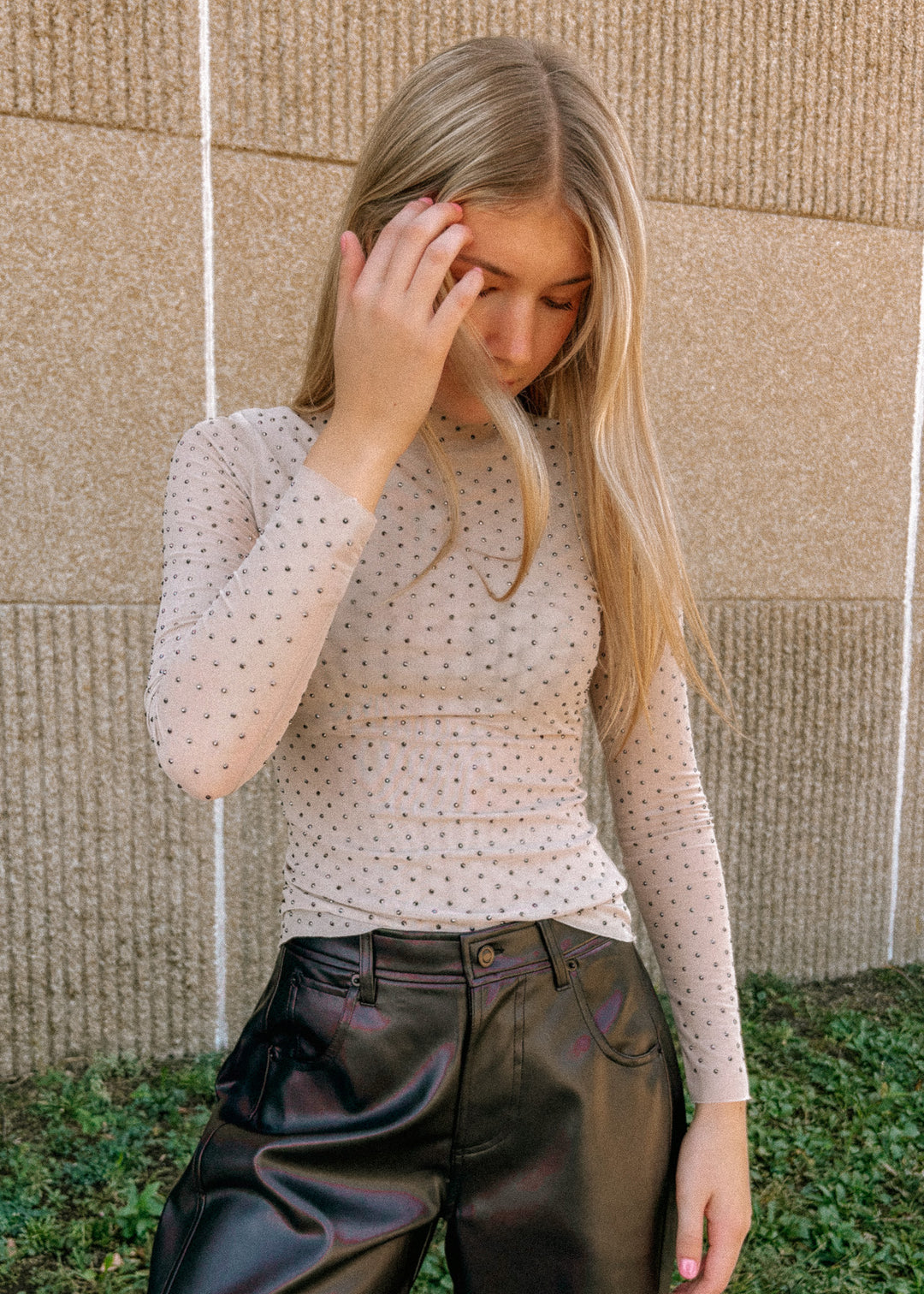 Woman wearing a mesh rhinestone top and black pants against a beige wall.