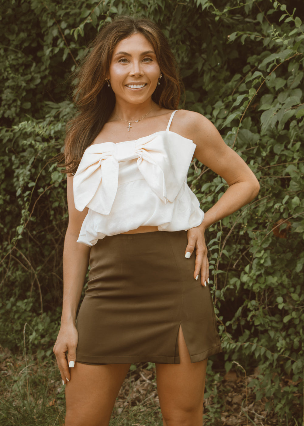 Woman wearing a white top with a bow and a green skirt standing in front of greenery.