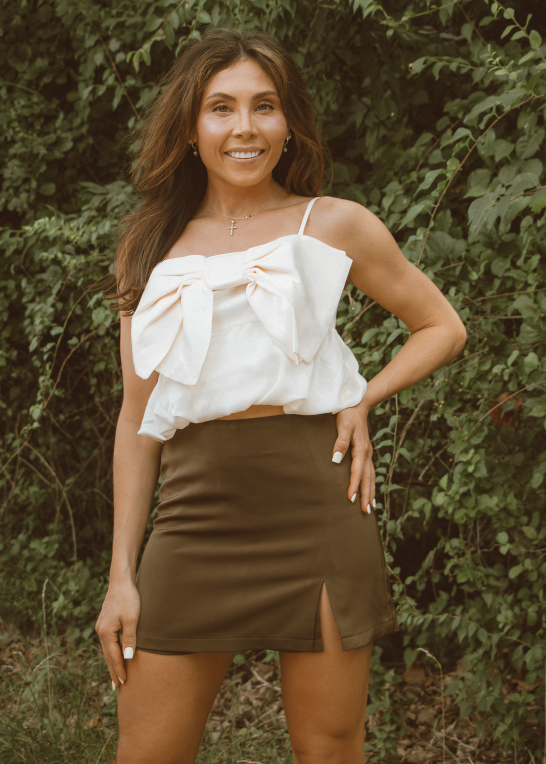 Woman wearing a white top with a bow and a green skirt standing in front of greenery.
