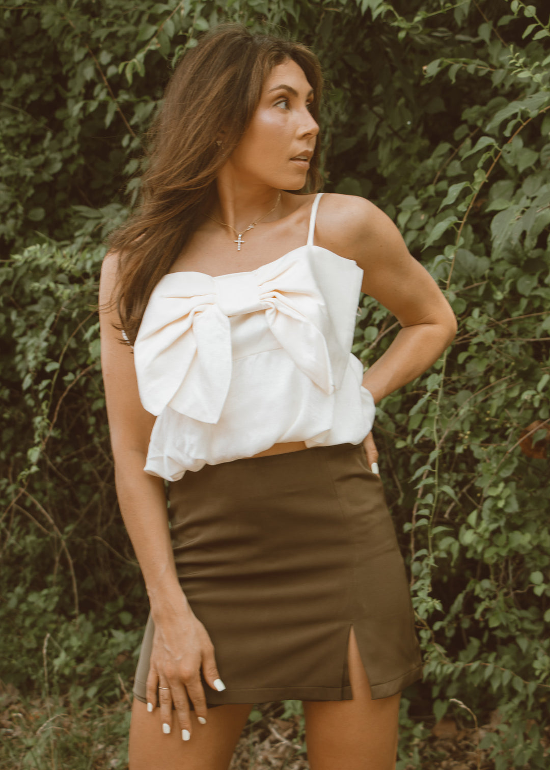 Woman wearing a white top with a bow and a brown skirt in front of green foliage