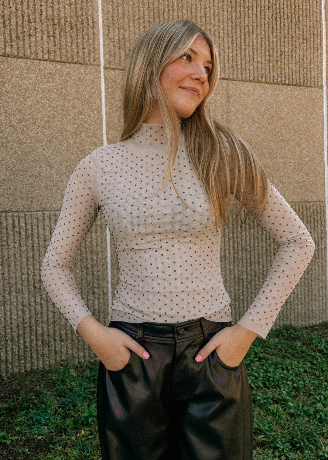 Woman wearing a long-sleeve top with rhinestones and black pants against a textured wall.