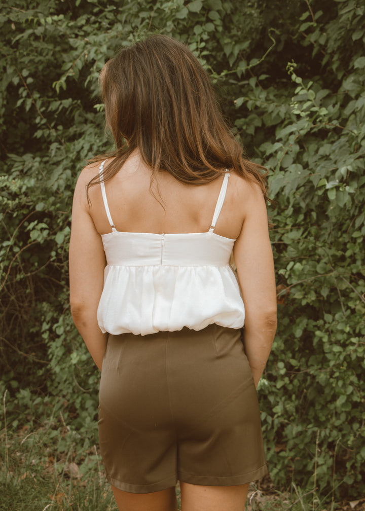Woman wearing a white top and brown shorts standing in front of green foliage
