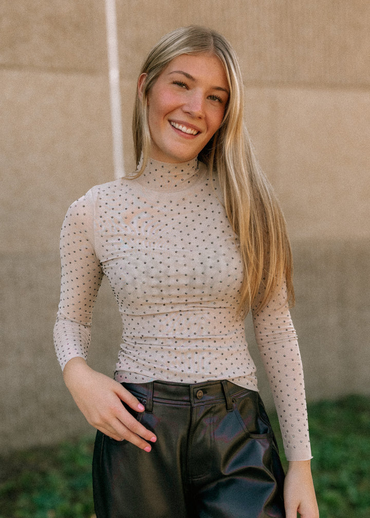 Woman wearing a mesh rhinestone top and black pants against a beige wall.