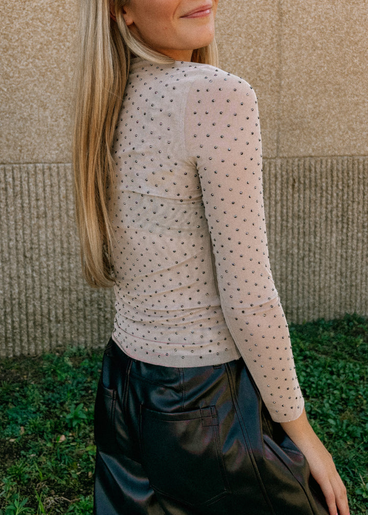 Woman wearing a beige long-sleeve top with rhinestones against a textured wall.