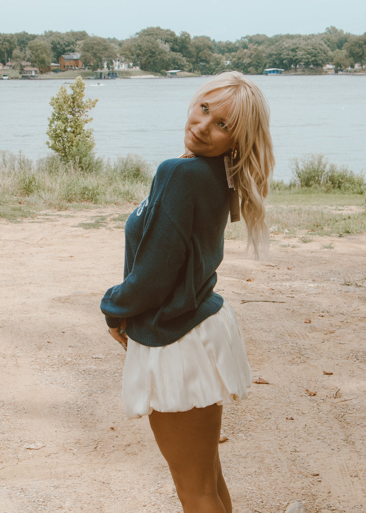 Woman standing by a lake wearing a green sweater and white skirt.