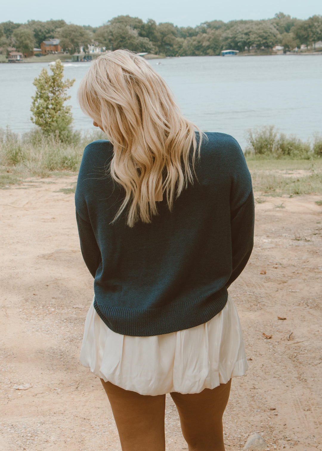 Woman standing by a lake wearing a dark green sweater and white skirt.