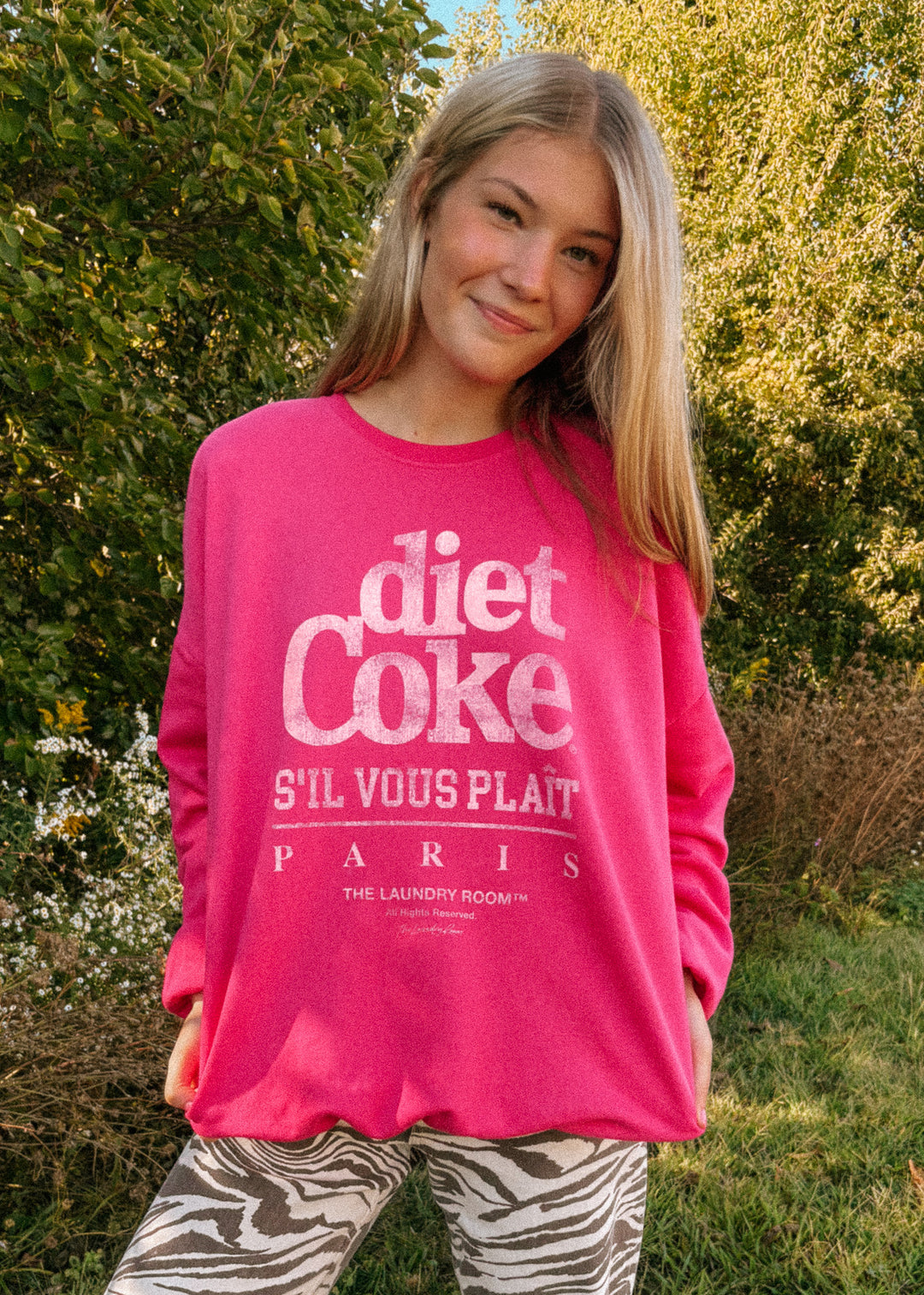 Person wearing a pink 'Diet Coke S'il Vous Plait Paris' sweatshirt outdoors.