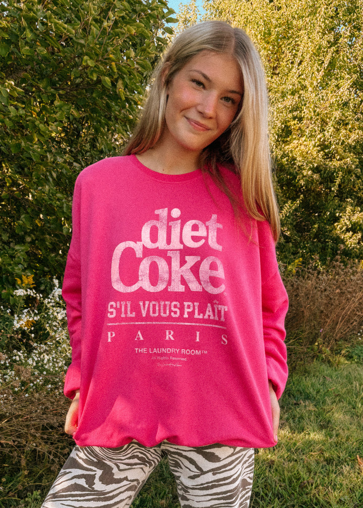 Person wearing a pink 'Diet Coke S'il Vous Plait Paris' sweatshirt outdoors.