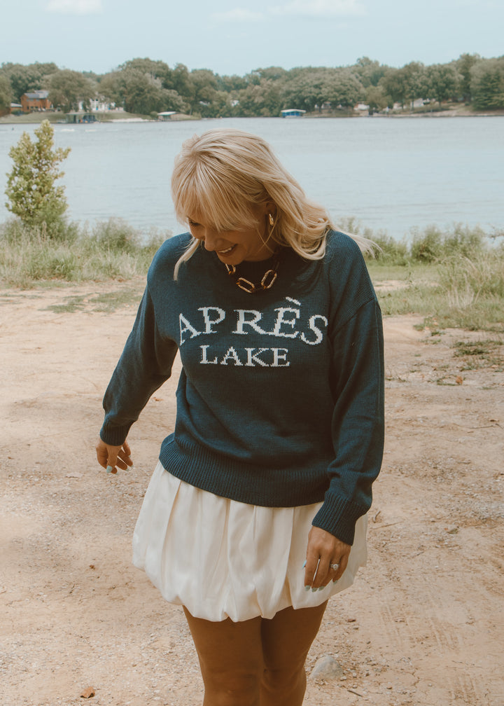 Woman wearing a sweatshirt with 'APRèS LAKE' text by a lake.
