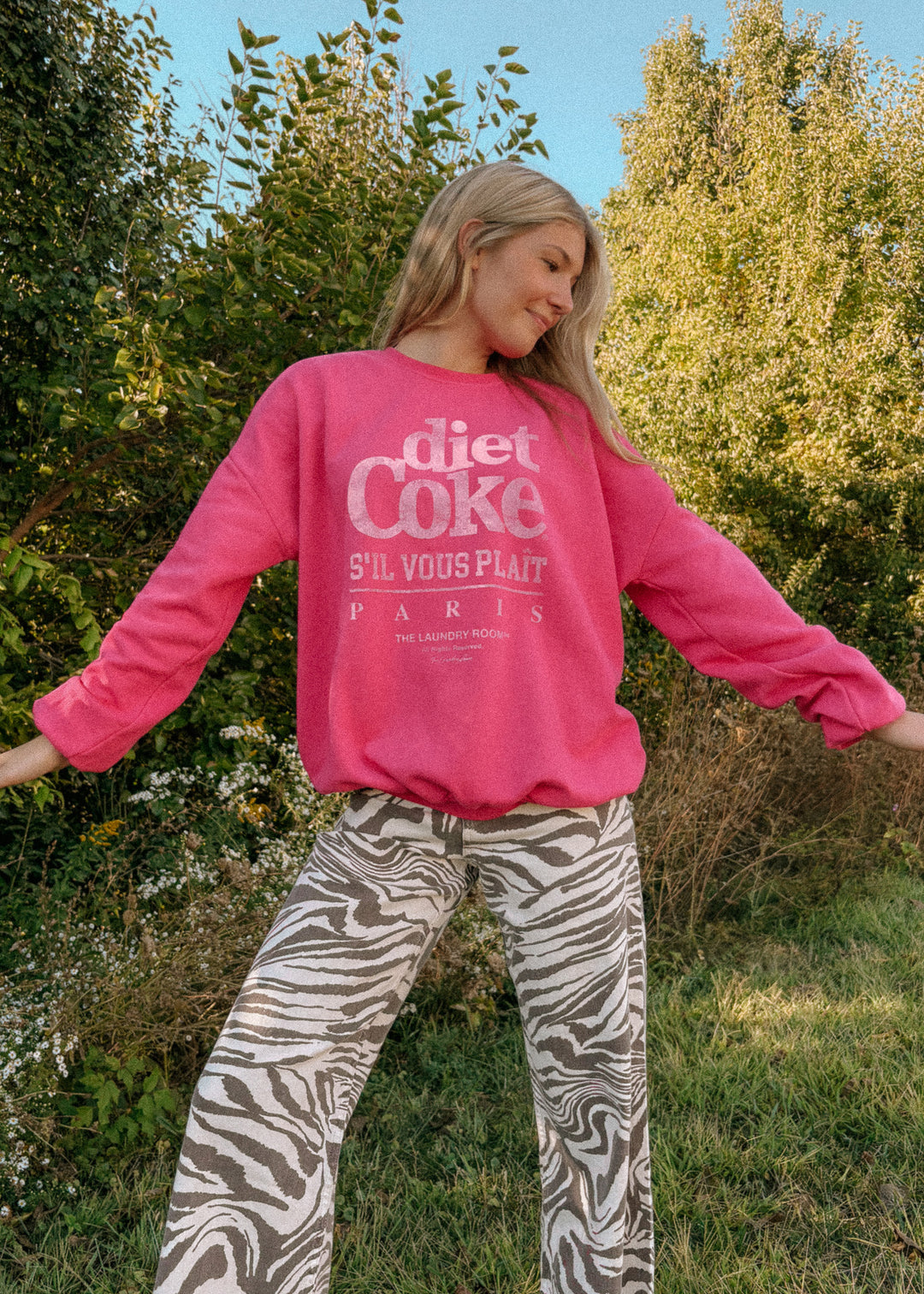 Person wearing a pink sweatshirt with 'diet Coke' text, standing outdoors with trees in the background
