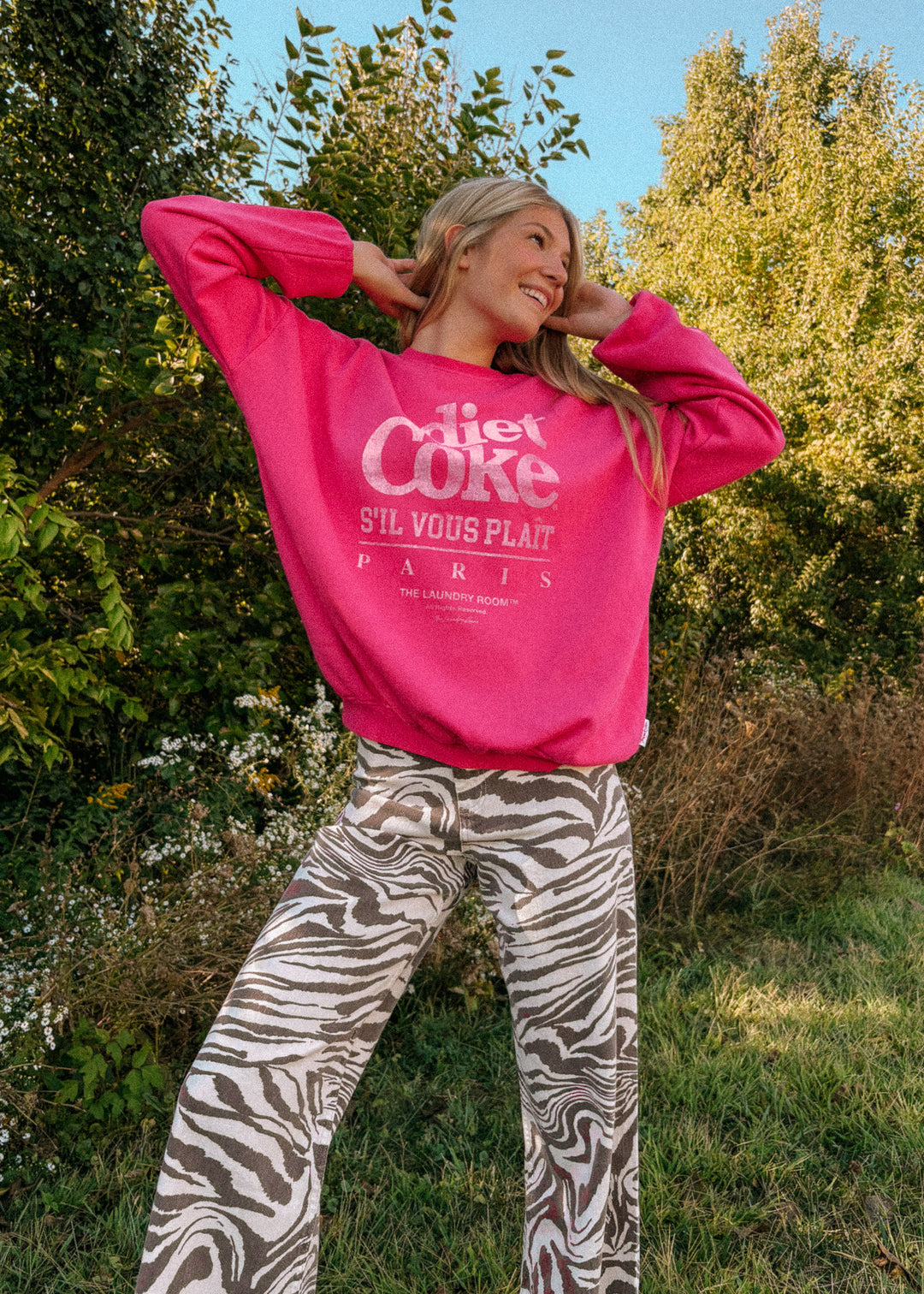 Person wearing a bright pink sweatshirt with text, standing outdoors with trees and grass in the background.