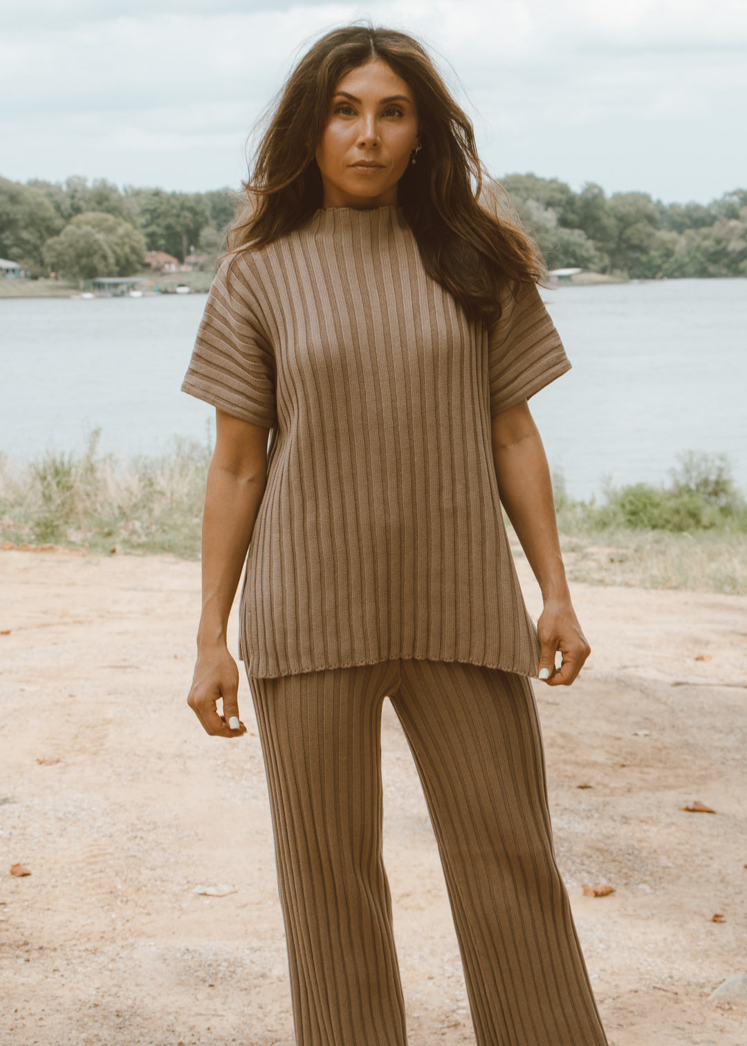 Woman wearing a brown ribbed sweater and pants standing by a lake.
