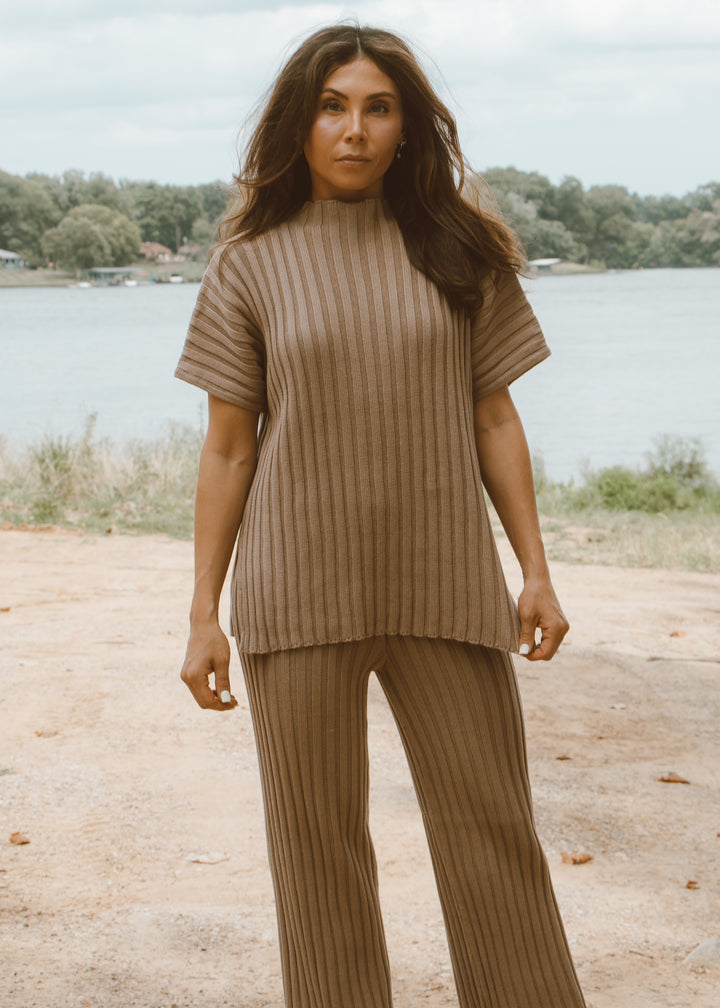 Woman wearing a brown ribbed sweater and pants standing by a lake.