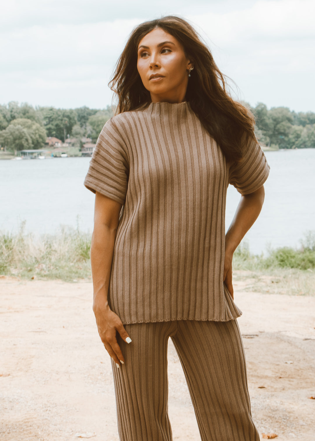 Woman wearing a beige ribbed sweater and pants standing by a lake.