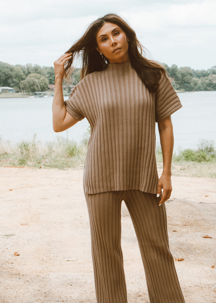 Woman wearing a brown ribbed sweater and pants by a lake