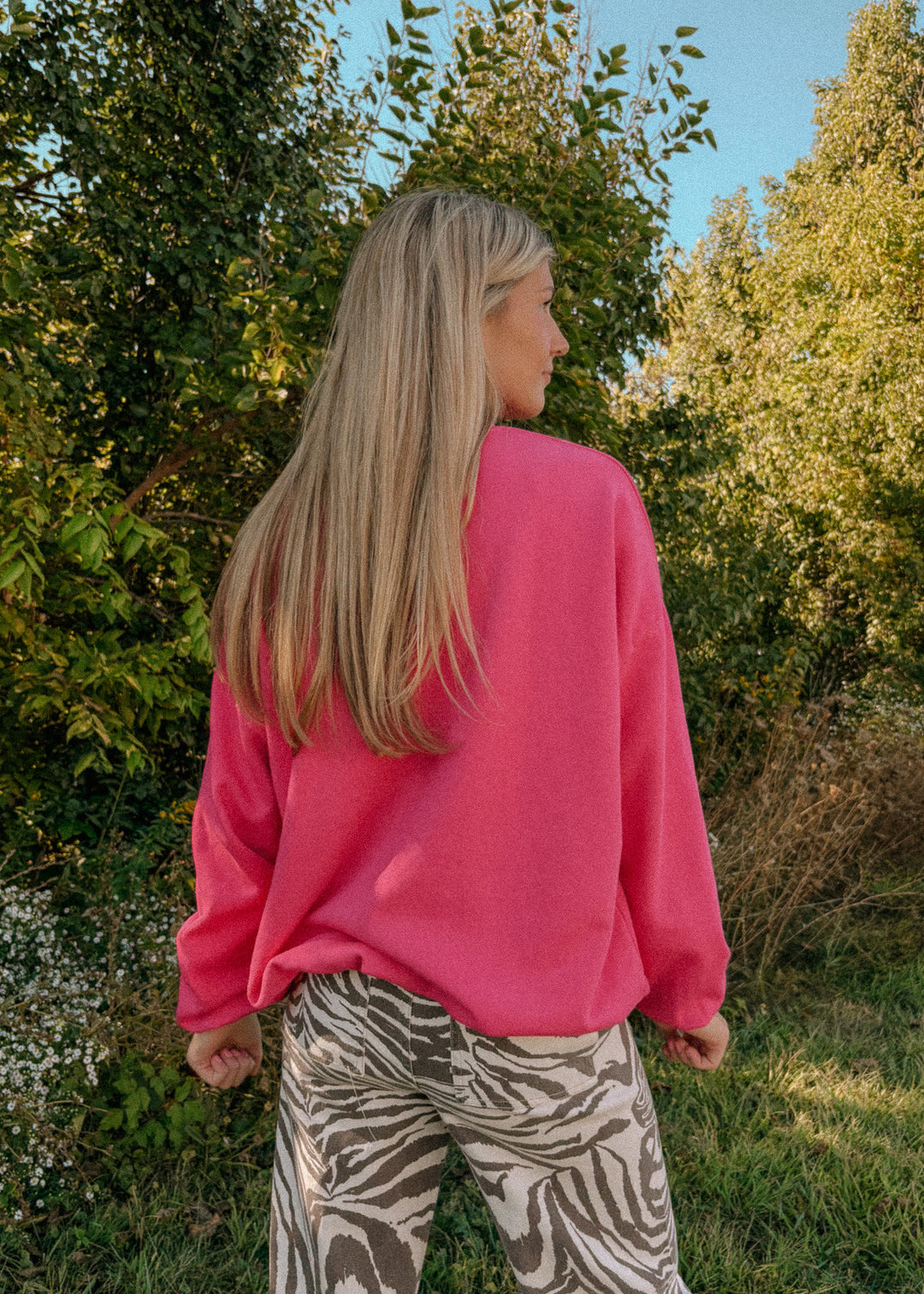 Person wearing a bright pink sweater and patterned pants standing in a natural setting with trees and greenery.