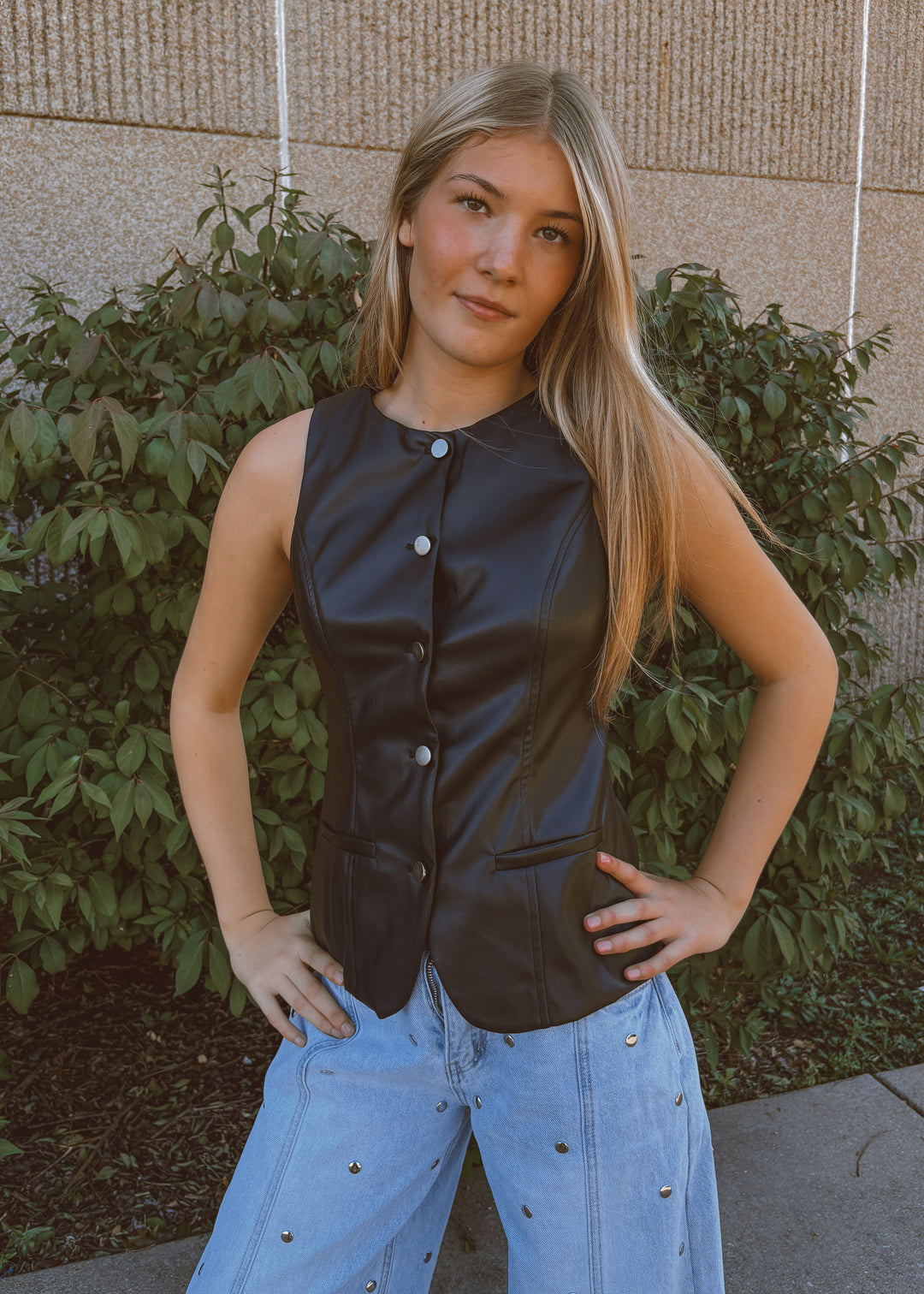Woman wearing a black sleeveless vest and blue jeans standing outdoors against a brick wall.