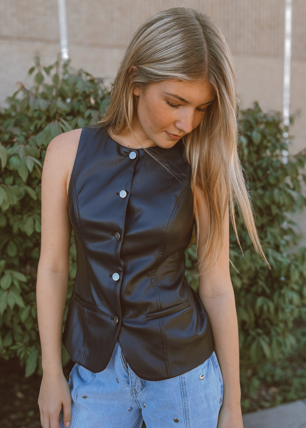 Woman wearing a black vest with buttons, standing outdoors against a green bush background.
