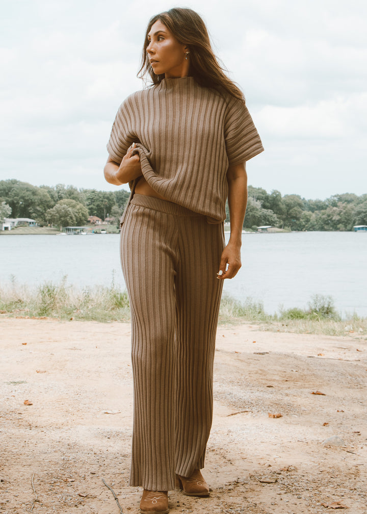 Woman wearing a brown ribbed sweater and pants standing by a lake.