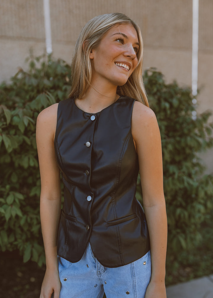 Woman wearing a black sleeveless top with buttons, standing outdoors with greenery in the background.
