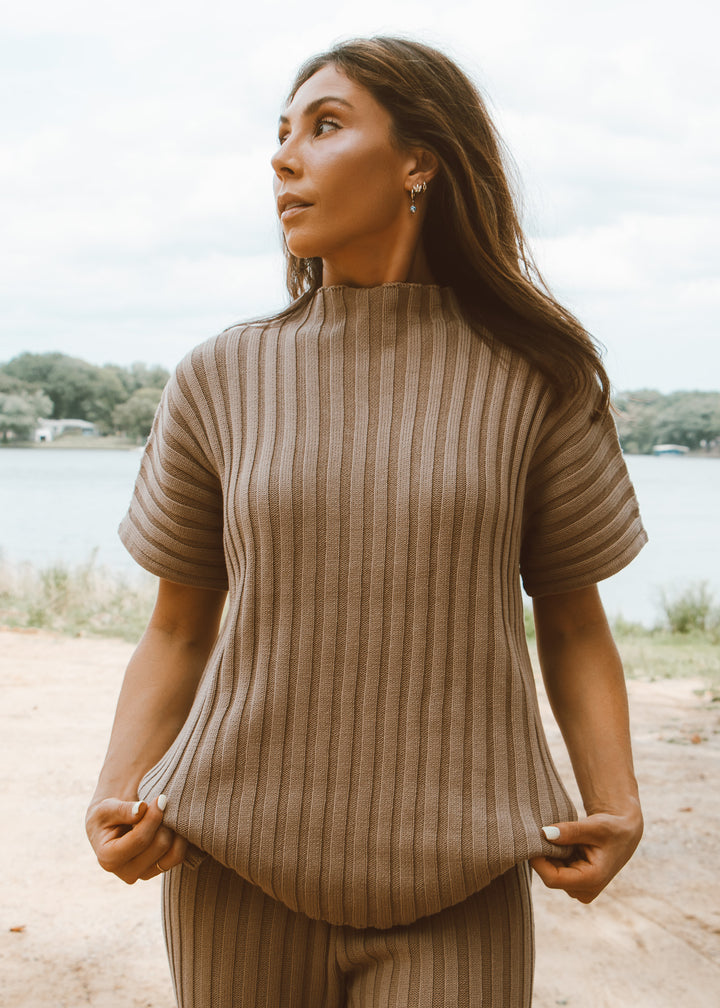 Woman wearing a beige ribbed sweater standing outdoors by a lake.