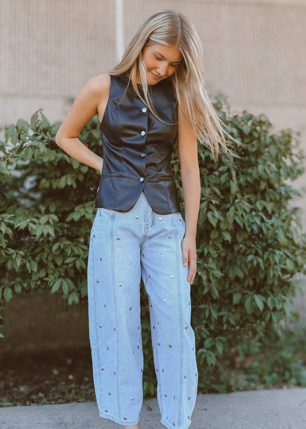 Woman wearing a black leather vest and light blue jeans standing in front of greenery.