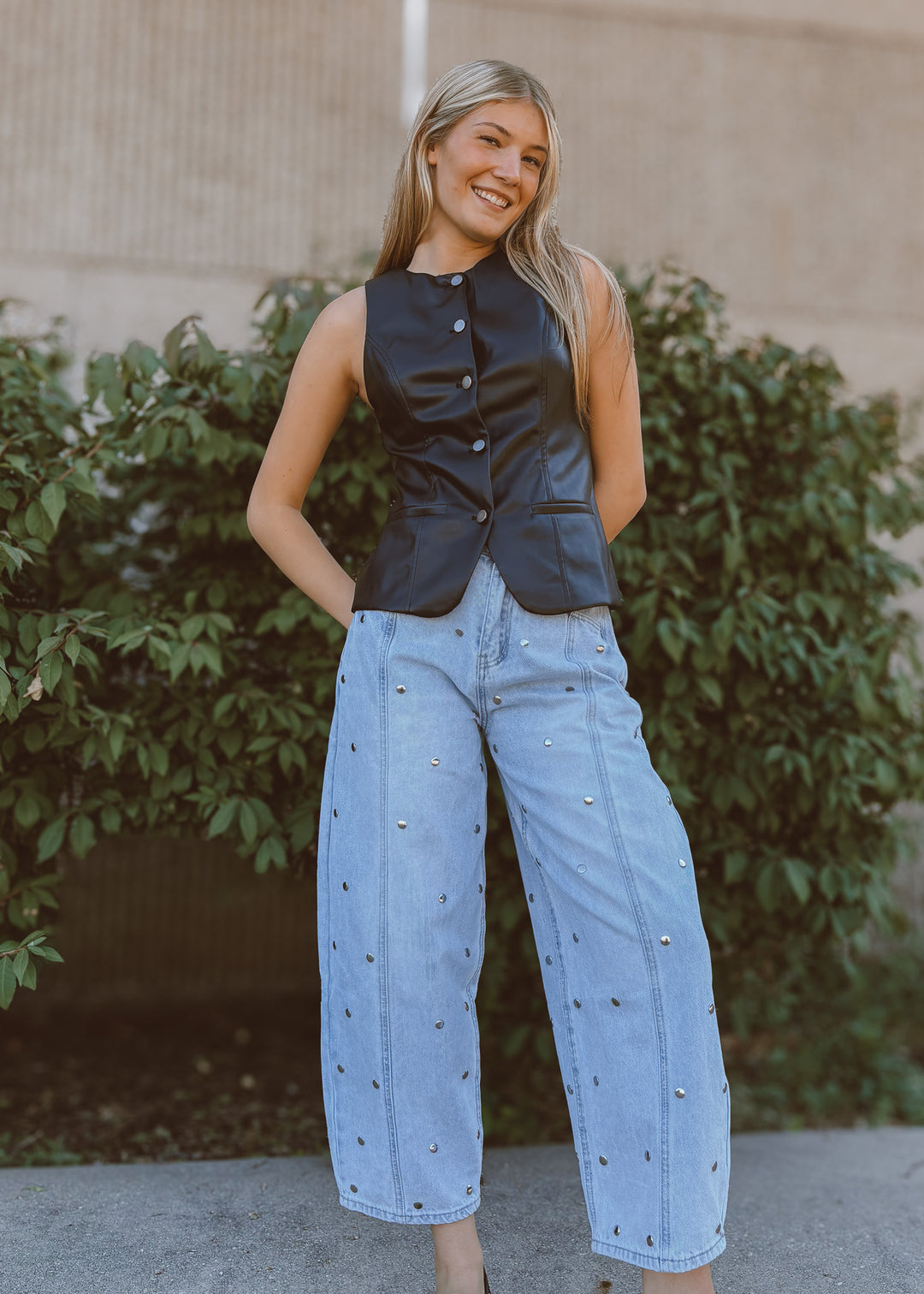 Woman wearing a black leather vest and light blue barrel jeans standing outdoors with greenery in the background