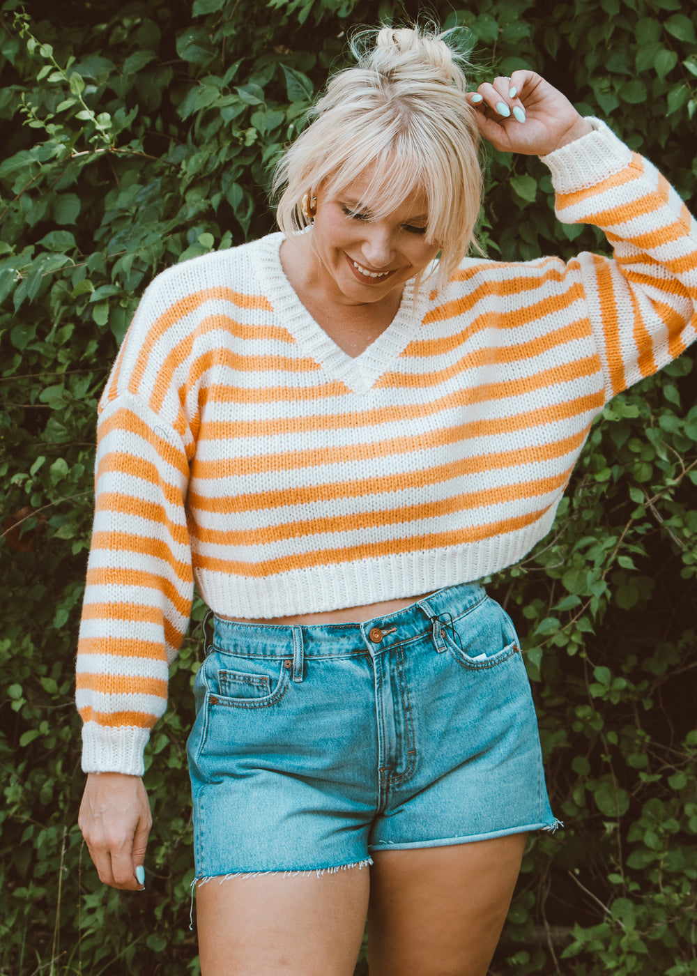 Woman wearing a striped sweater and denim shorts standing in front of green foliage