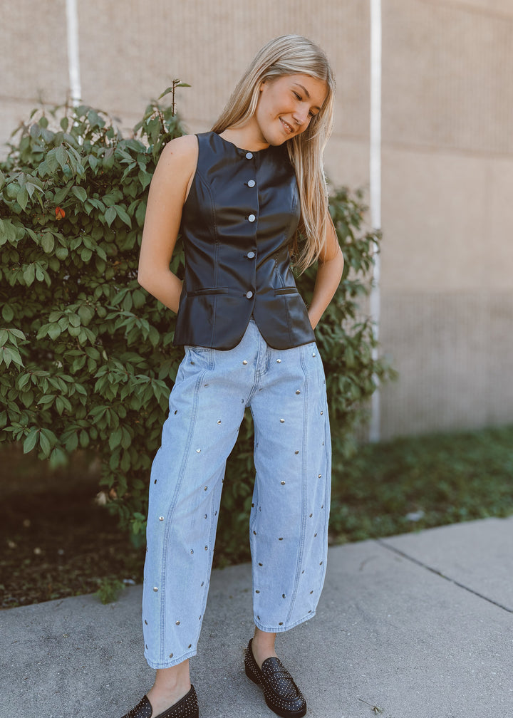 Woman wearing a leather vest and light blue pants standing outdoors.