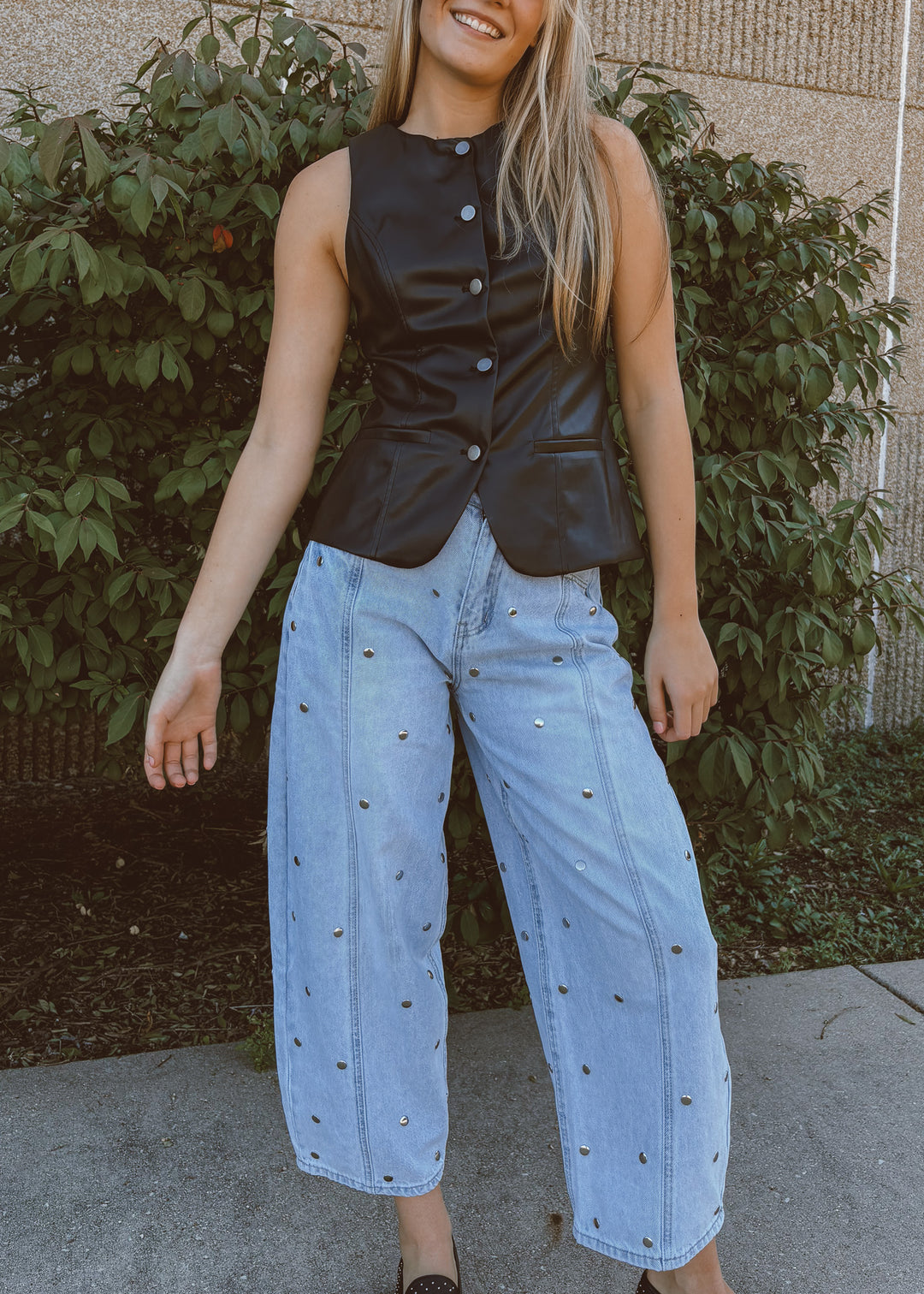 Woman wearing a black vest and light blue jeans standing outdoors.