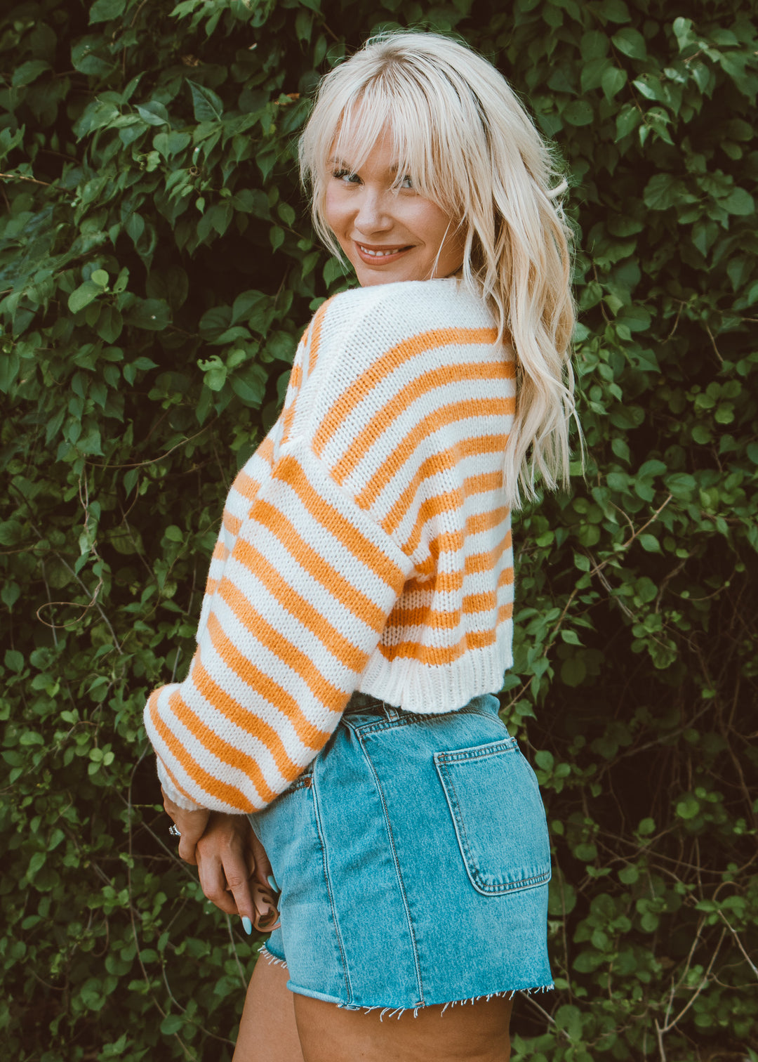 Person wearing a striped sweater and denim shorts standing against a green leafy background