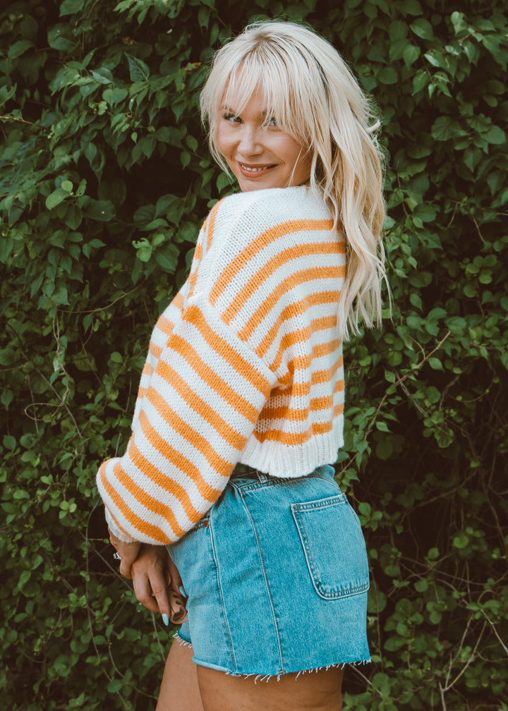 Person wearing a striped sweater and denim shorts standing against a green leafy background