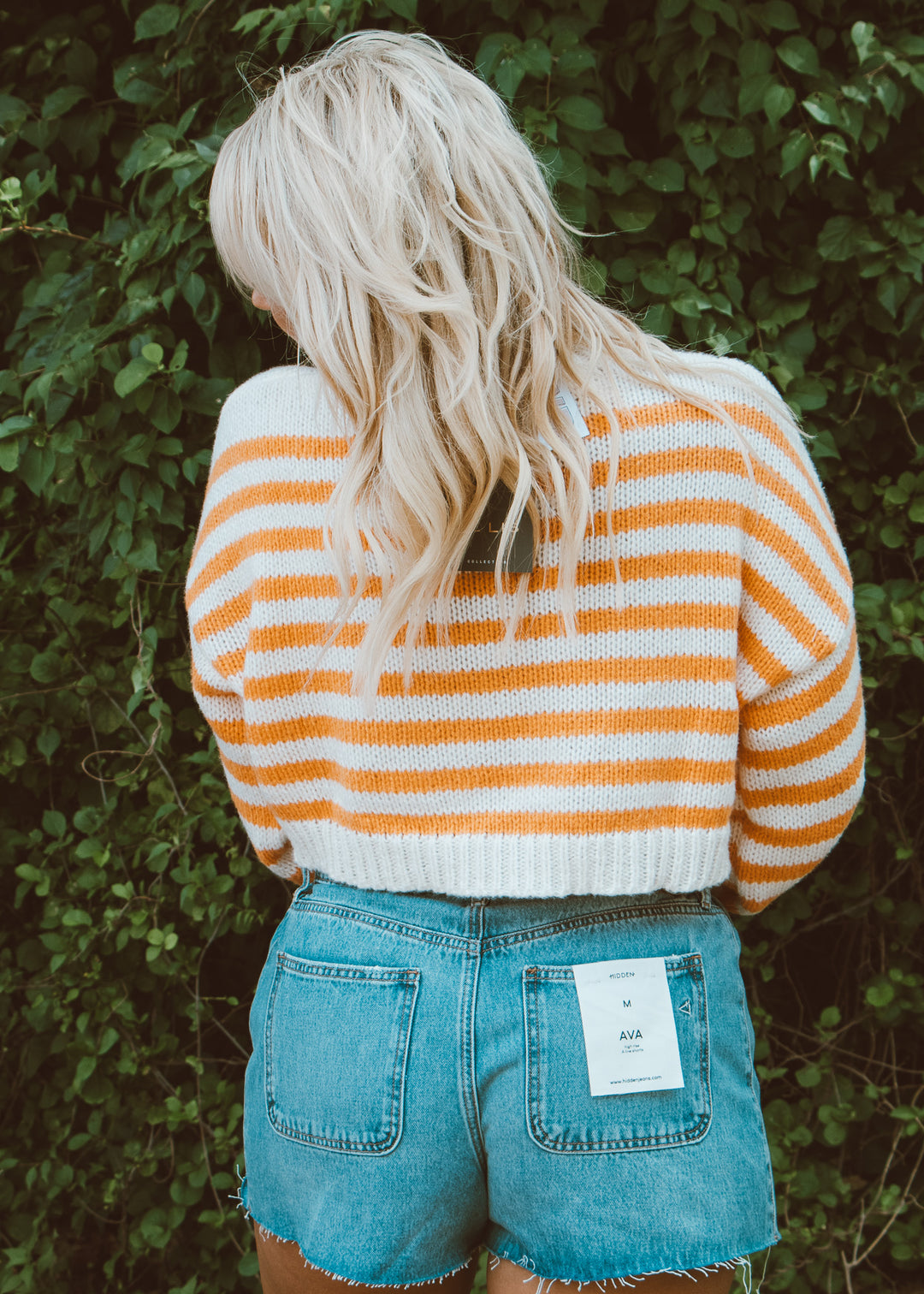 Person wearing an orange and white striped sweater and blue denim shorts with a brand tag, standing against a green leafy background.