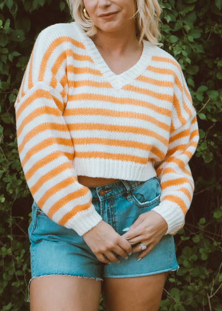 Person wearing an orange and white striped sweater with denim shorts against a green leafy background