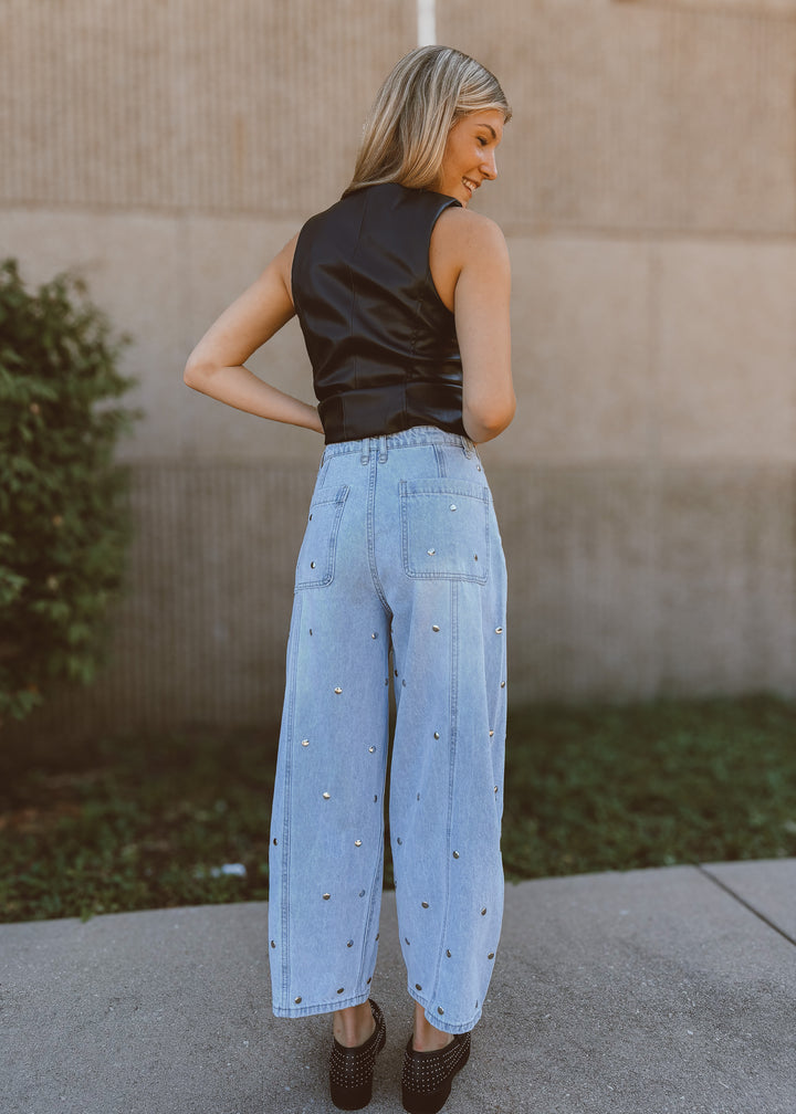 Woman wearing a black leather vest and light blue jeans with rhinestone details, standing against a beige wall.