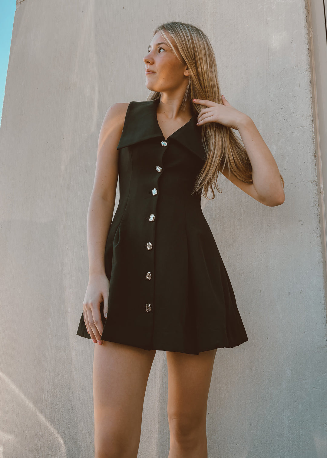 Woman wearing a black sleeveless dress with buttons against a plain wall.