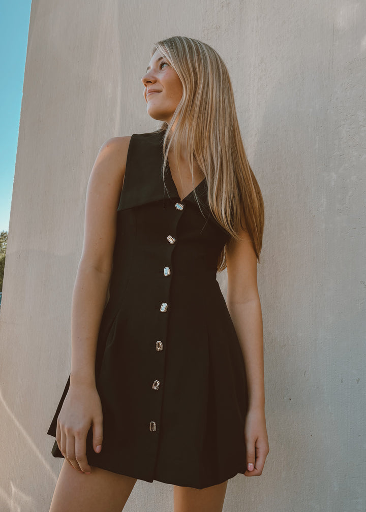 Woman wearing a black dress with gold buttons against a light-colored wall.