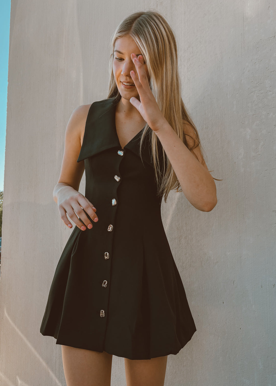 Woman wearing a black dress with gold buttons against a light-colored wall.
