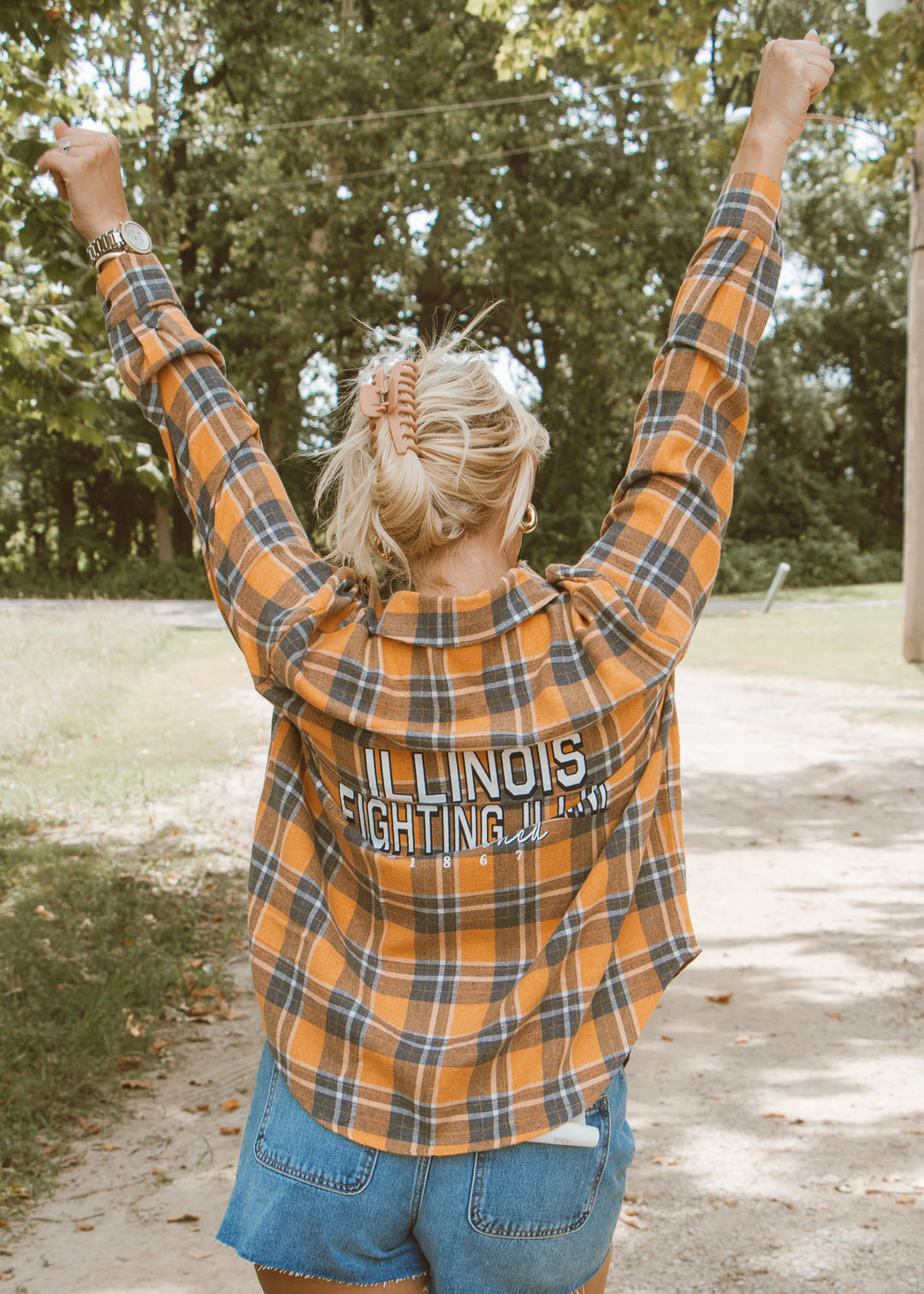 Person wearing an 'Illinois Fighting Illini' plaid shirt with arms raised outdoors.
