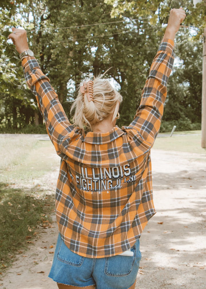 Person wearing an 'Illinois Fighting Illini' plaid shirt with arms raised outdoors.