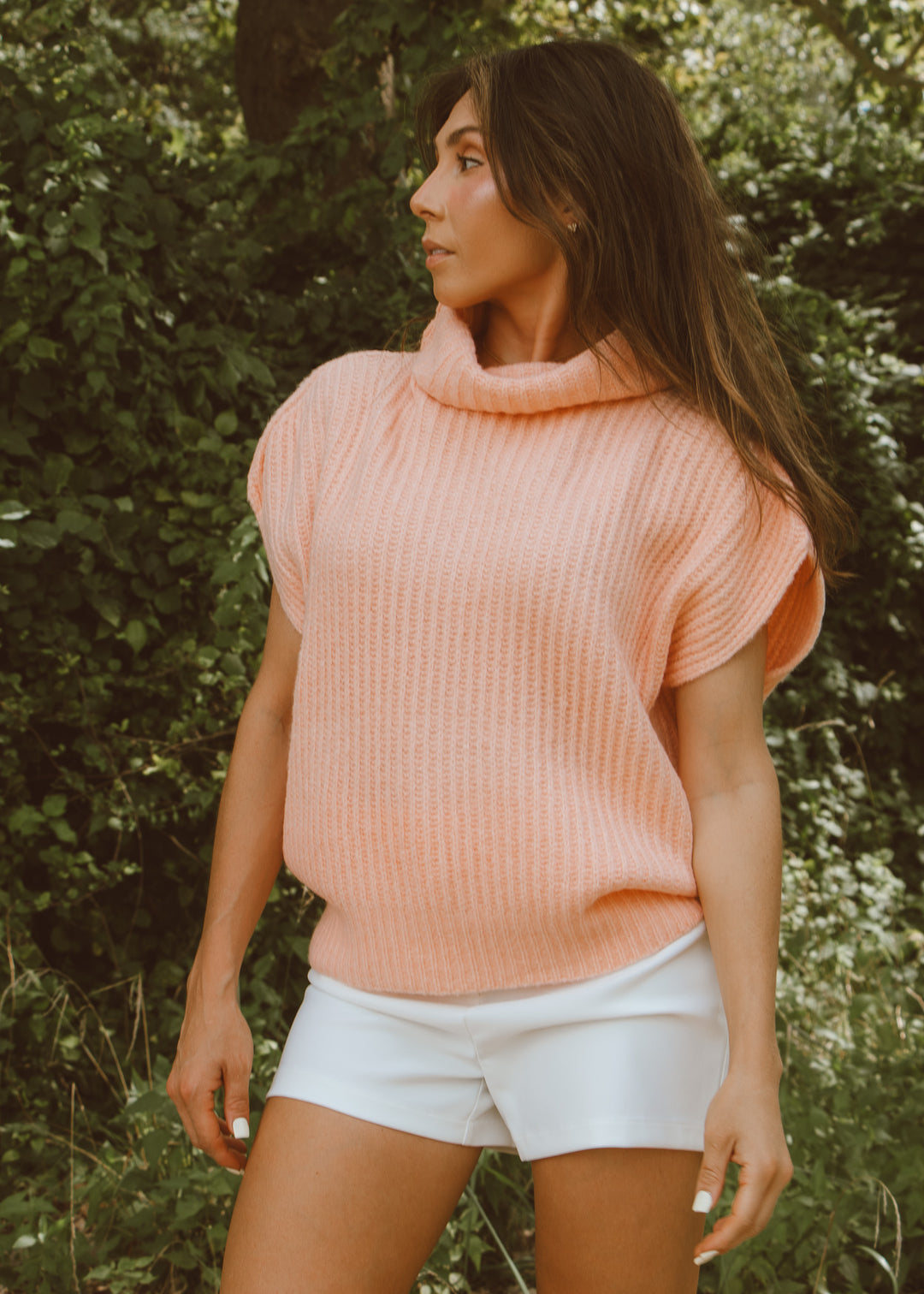 Woman wearing a pink sweater and white shorts standing in front of green foliage