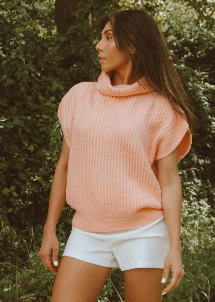 Woman wearing a pink sweater and white shorts standing in front of green foliage