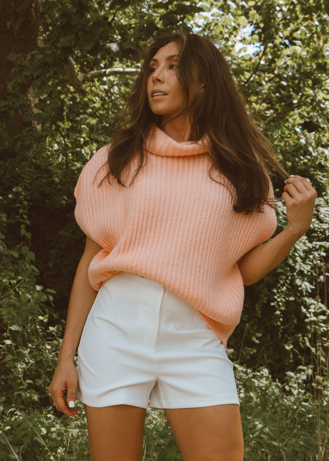 Woman wearing a pink sweater and white shorts standing in a forest.