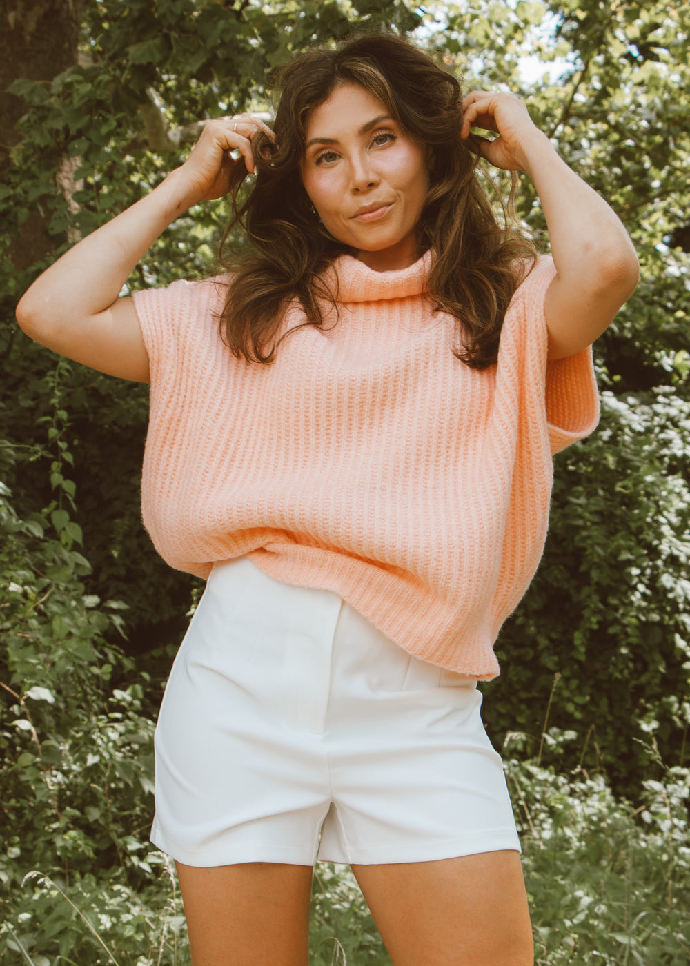 Woman wearing a pink sweater vest and white shorts standing in a natural setting with greenery.