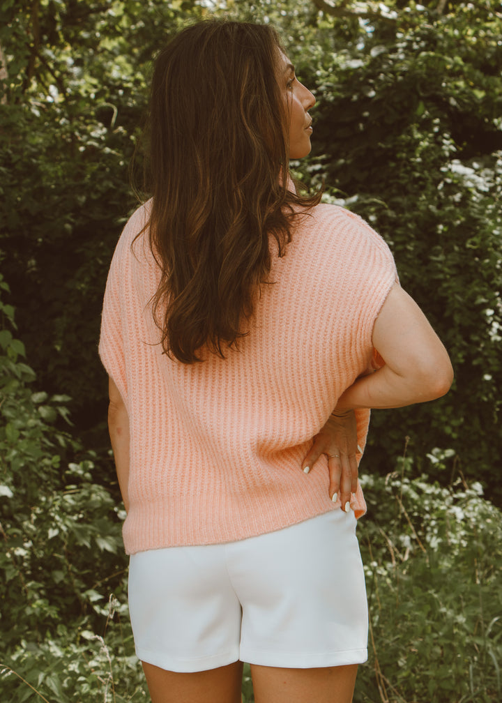 Woman wearing a pink sleeveless top and white shorts standing in a natural setting with greenery.